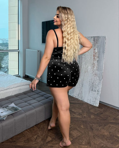 A woman with long blonde wavy hair stands in a bedroom wearing a black sleeveless romper with white polka dots, photographed from behind.