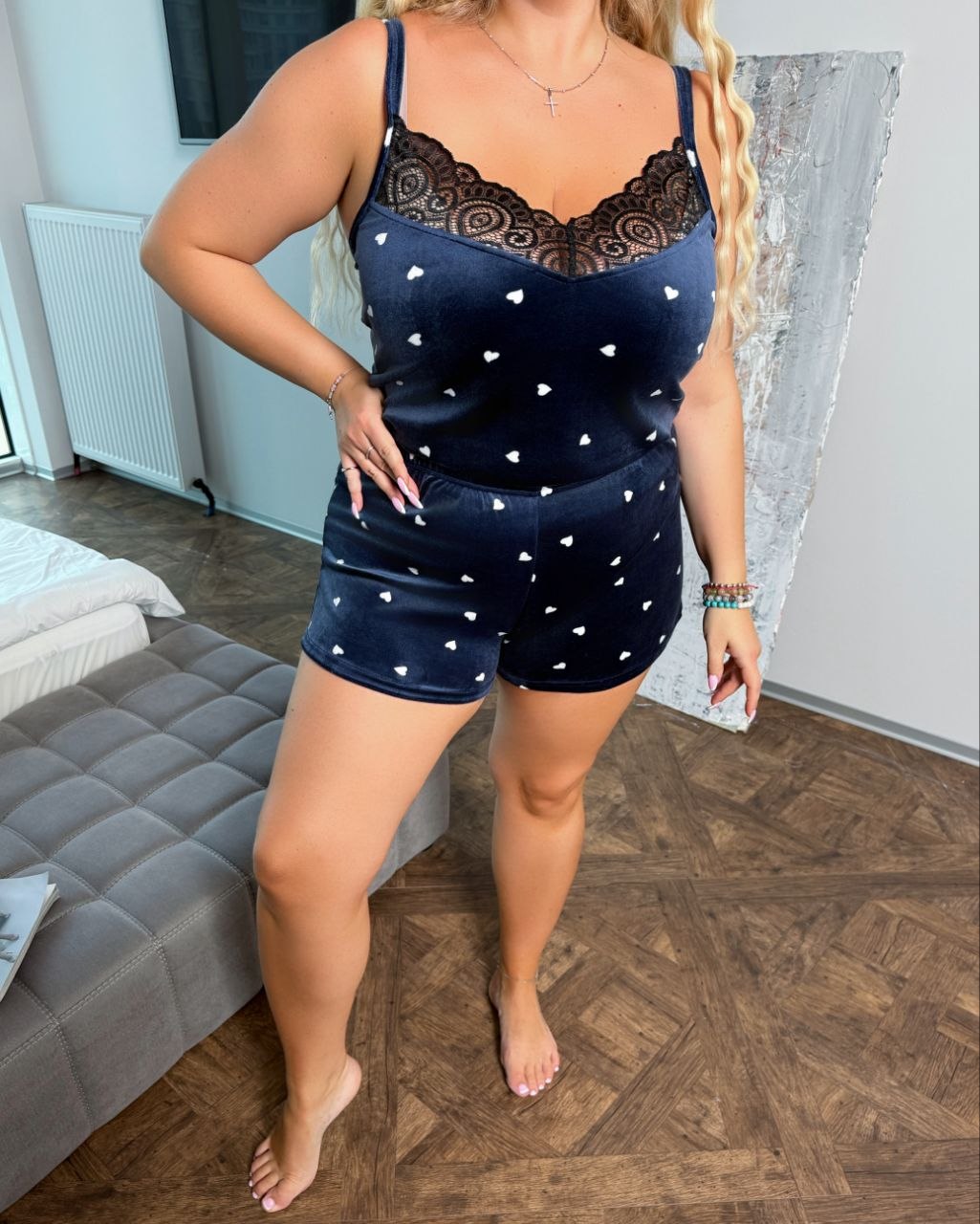 A woman modeling a navy blue velvet two-piece pajama set with white hearts and black lace trim, standing in a bedroom setting.