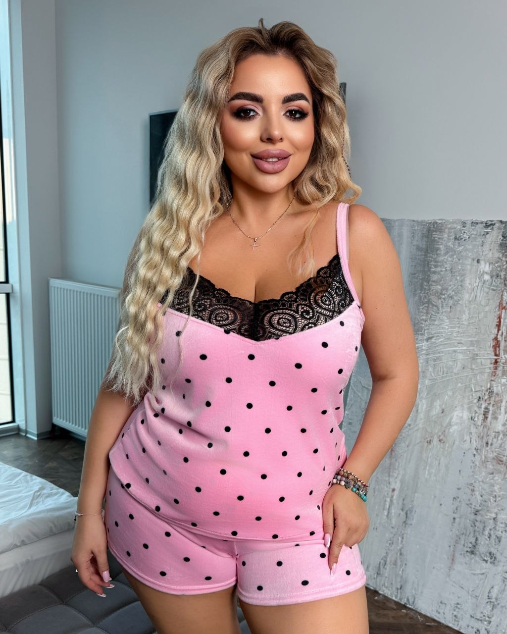 A woman modeling a pink two-piece pajama set with black polka dots and a black lace-trimmed neckline, standing indoors against a gray wall.