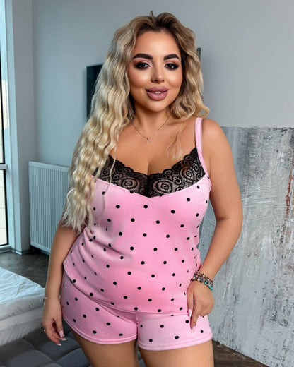 A woman modeling a pink two-piece pajama set with black polka dots and a black lace-trimmed neckline, standing indoors against a gray wall.