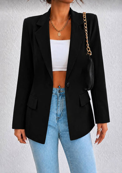 A woman wearing a black long-sleeve blazer over a white crop top and light blue jeans, with a black shoulder bag featuring a gold chain strap.