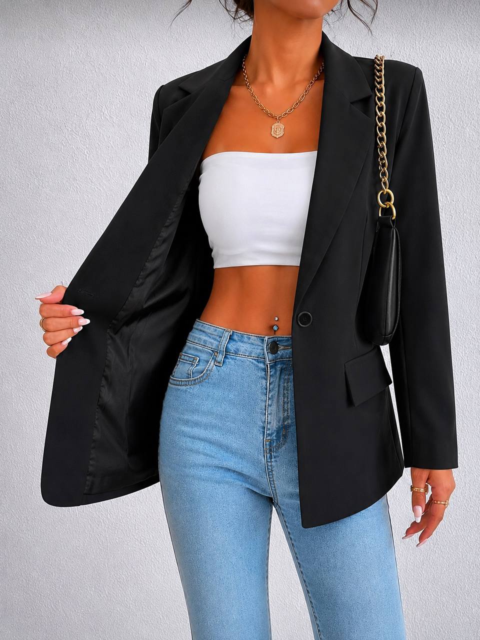 A woman wearing a black blazer over a white tube top and light blue jeans, holding the blazer open. She is carrying a black shoulder bag with a gold chain strap and wearing a gold necklace. The background is a textured light gray wall.