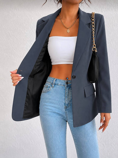 A woman wearing a dark slate blue blazer over a white tube top and light blue jeans, carrying a black shoulder bag with a gold chain strap.