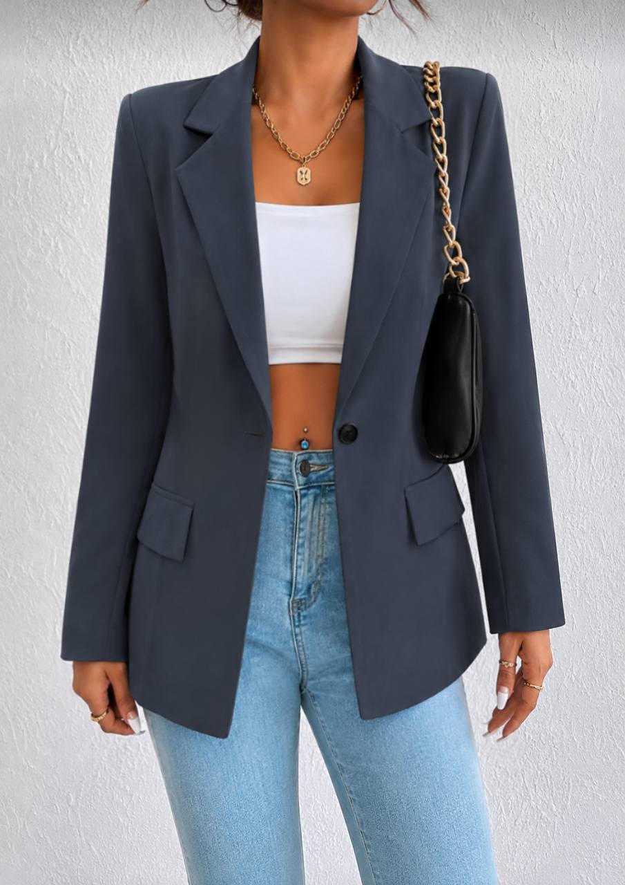 A woman wearing a dark slate blue blazer over a white crop top and light blue jeans, carrying a black shoulder bag with a gold chain strap against a textured white wall.