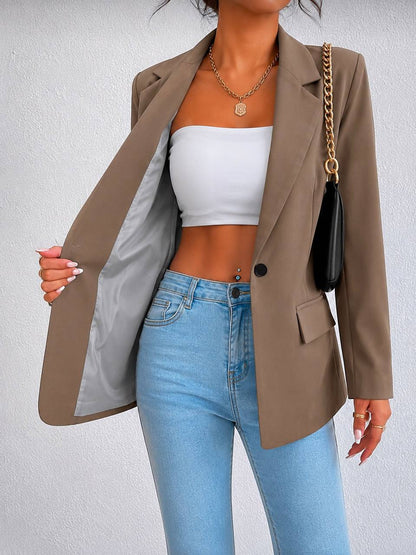 A woman wearing a taupe brown blazer over a white bandeau top and light blue jeans, holding the blazer open to show the gray lining. She is also wearing a black shoulder bag with a gold chain strap and gold necklaces.