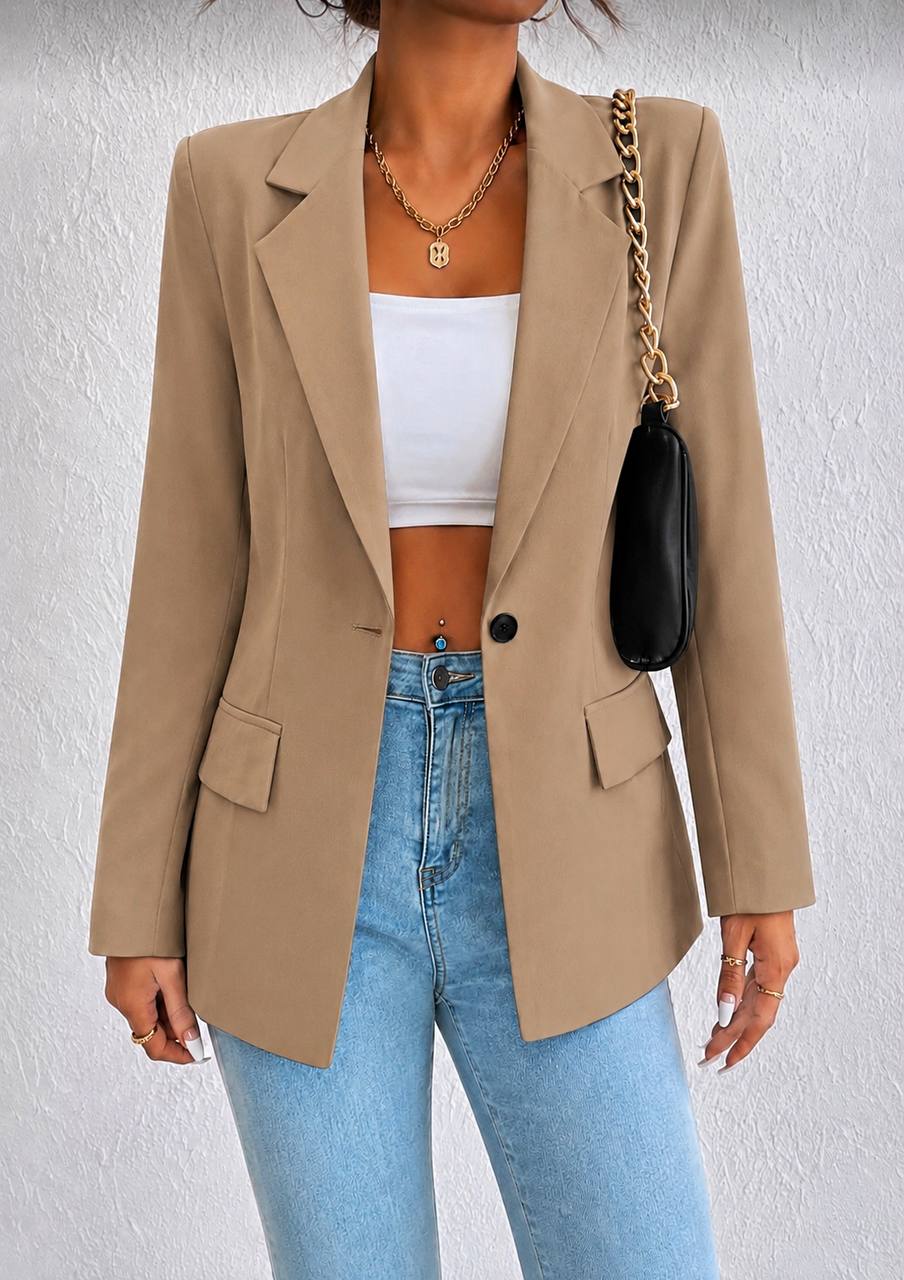 A woman wearing a beige blazer over a white crop top and light blue jeans, carrying a black shoulder bag with a gold chain strap against a textured white wall.