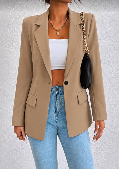 A woman wearing a beige blazer over a white crop top and light blue jeans, carrying a black shoulder bag with a gold chain strap against a textured white wall.