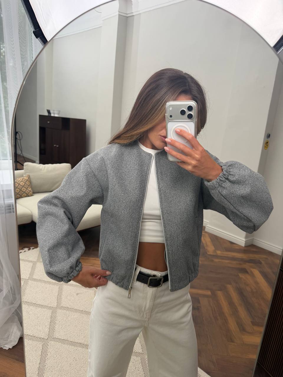 A woman takes a mirror selfie wearing a gray zip-up bomber jacket over a white crop top and white pants in a home setting.