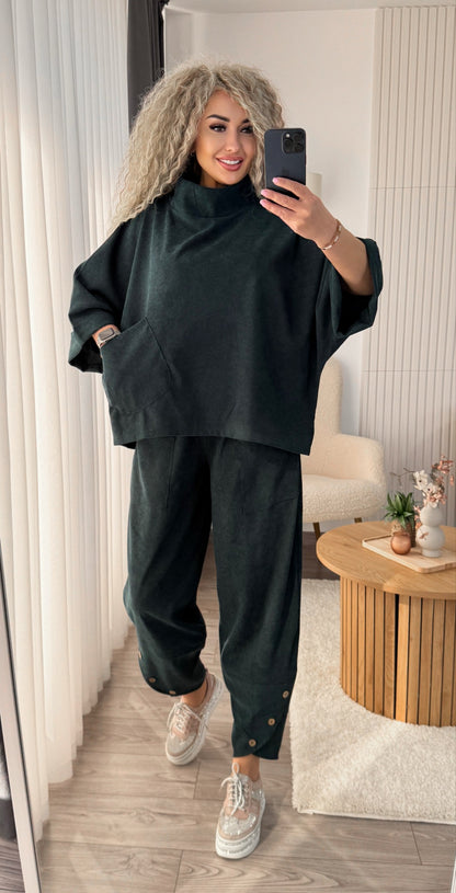 A woman takes a mirror selfie wearing a dark forest green two-piece lounge set consisting of a high-neck oversized sweatshirt and matching jogger pants, paired with white sneakers in a modern living room setting.