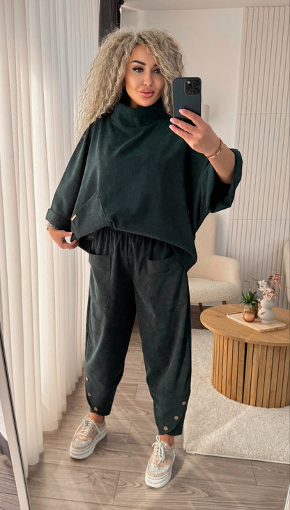 A woman takes a mirror selfie in a room wearing a dark forest green two-piece lounge set consisting of a turtleneck pullover and matching jogger pants. She is wearing white sneakers and holding a black smartphone.
