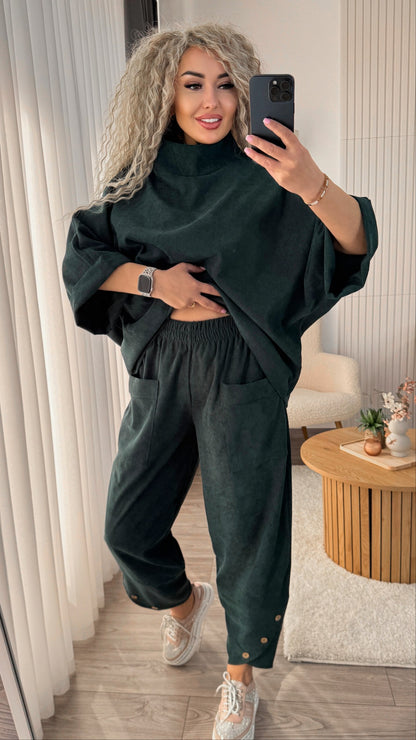 A woman takes a mirror selfie wearing a dark forest green two-piece lounge set consisting of a turtleneck top with wide sleeves and matching wide-leg pants with button details at the ankles, paired with white sneakers in a modern indoor setting.