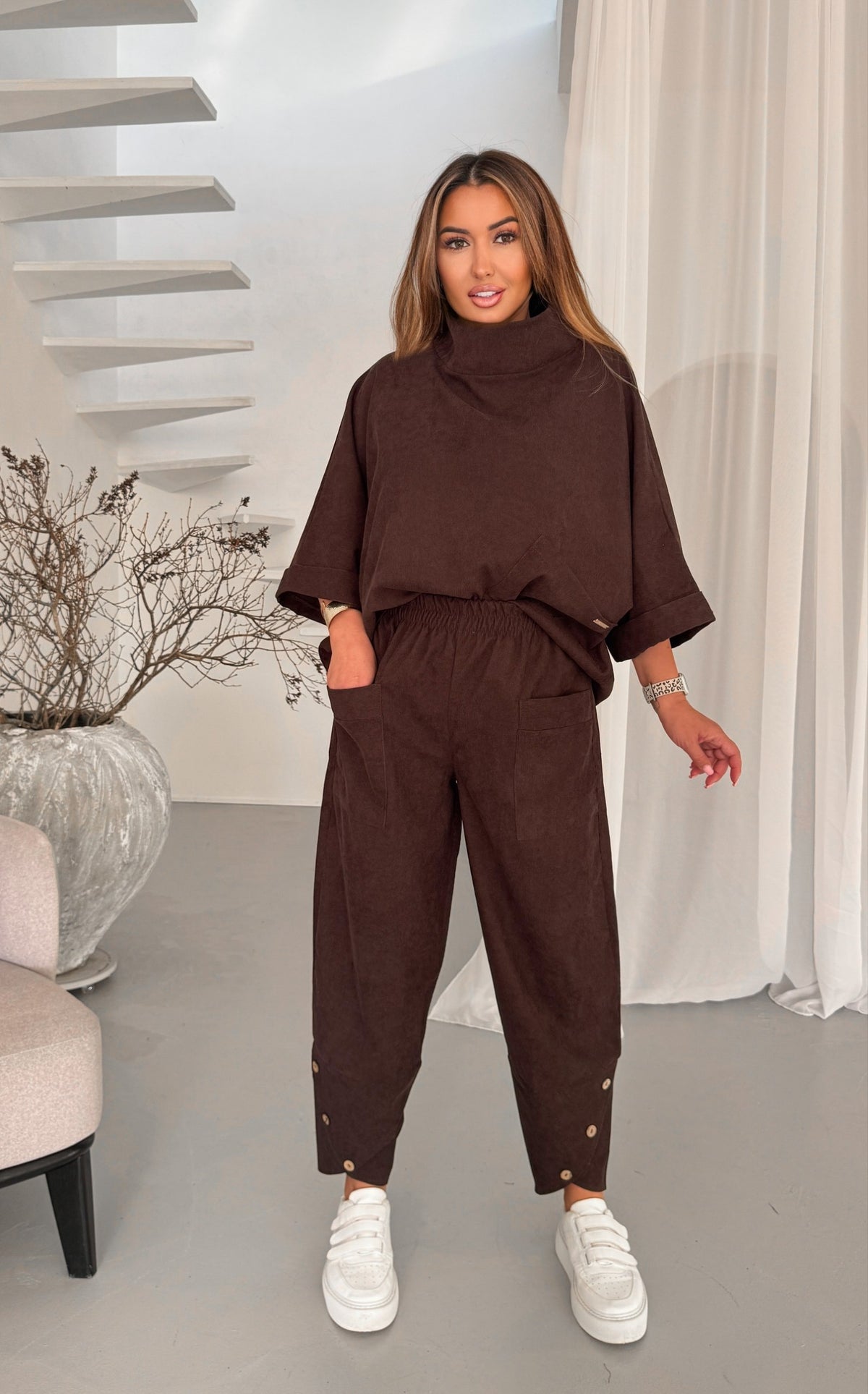 A woman modeling a dark brown two-piece lounge set consisting of a mock-neck short-sleeve top and matching drawstring pants with gold button details at the ankles, paired with white sneakers in a modern indoor setting.