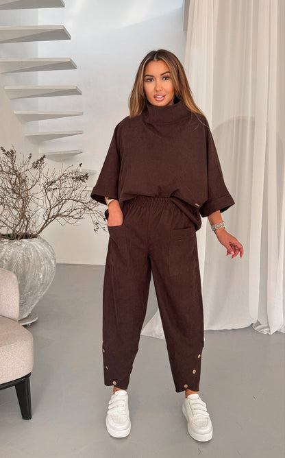 A woman modeling a dark brown two-piece lounge set consisting of a mock-neck short-sleeve top and matching drawstring pants with gold button details at the ankles, paired with white sneakers in a modern indoor setting.
