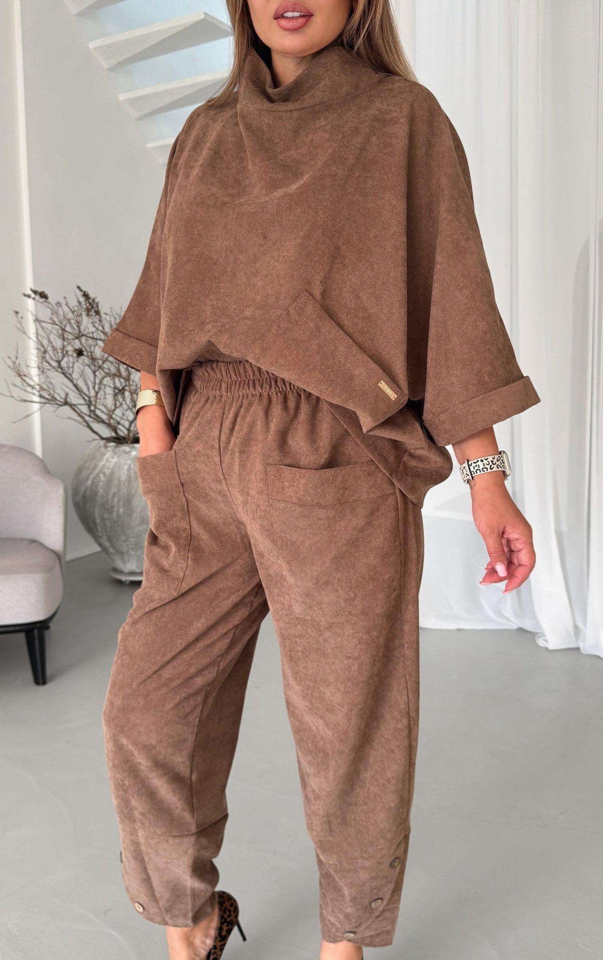A woman wearing a matching brown two-piece lounge set consisting of a high-neck, oversized short-sleeve top and matching elastic-waist pants with side pockets, shown in a bright indoor setting.