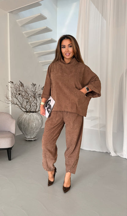 A woman modeling a matching brown lounge set consisting of a cropped, oversized long-sleeve top and wide-leg trousers, styled with brown pointed-toe heels in a modern, bright interior setting.