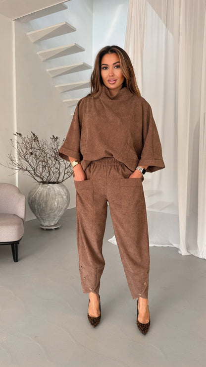 A woman modeling a matching brown corduroy lounge set consisting of a high-neck turtleneck top and relaxed-fit pants, standing in a modern, brightly lit interior with white walls, a staircase, and a gray floor.