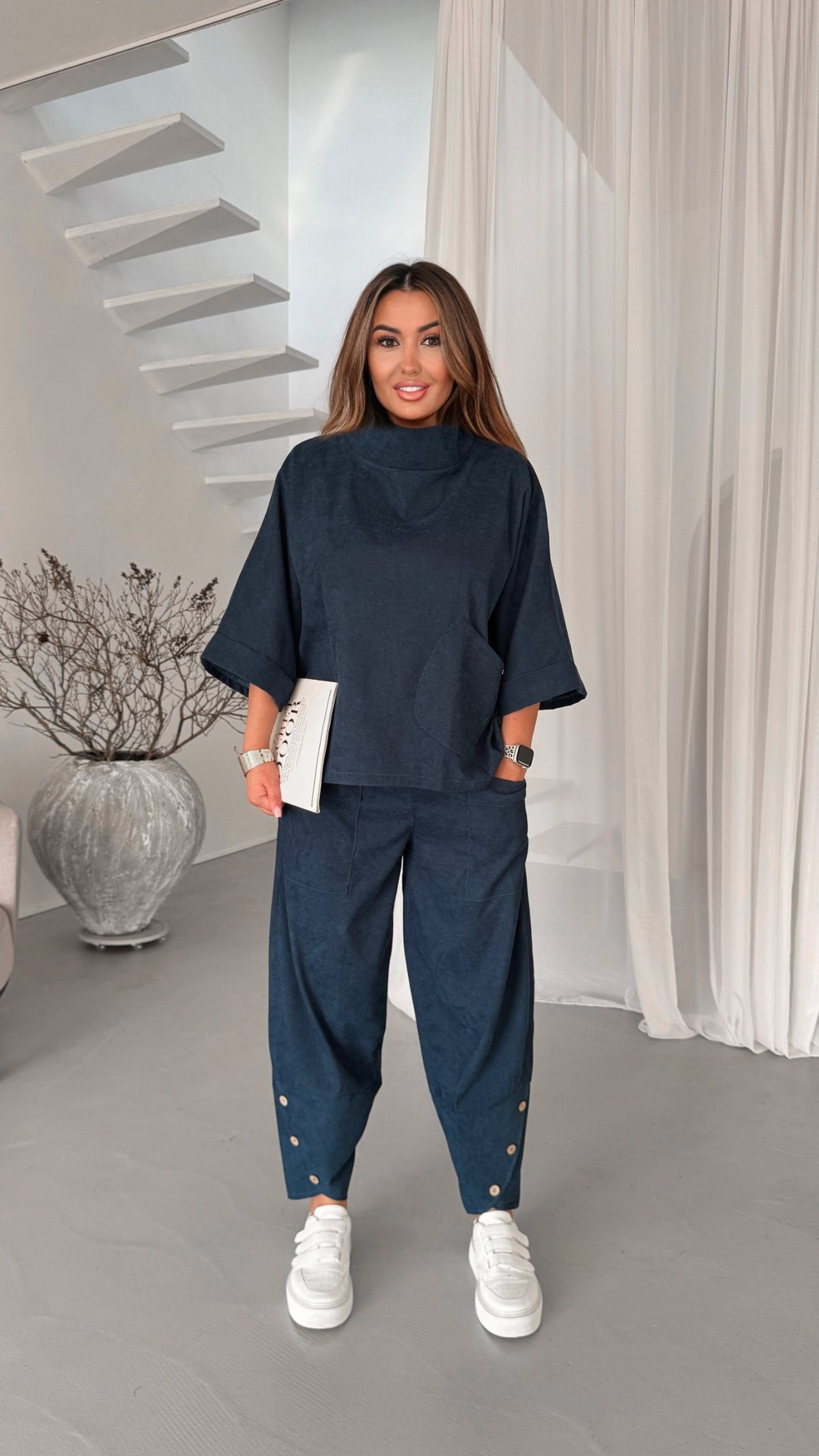 A woman modeling a matching navy blue lounge set consisting of a short-sleeve mock-neck top and relaxed-fit pants with gold button details at the ankles, paired with white sneakers in a modern interior setting.