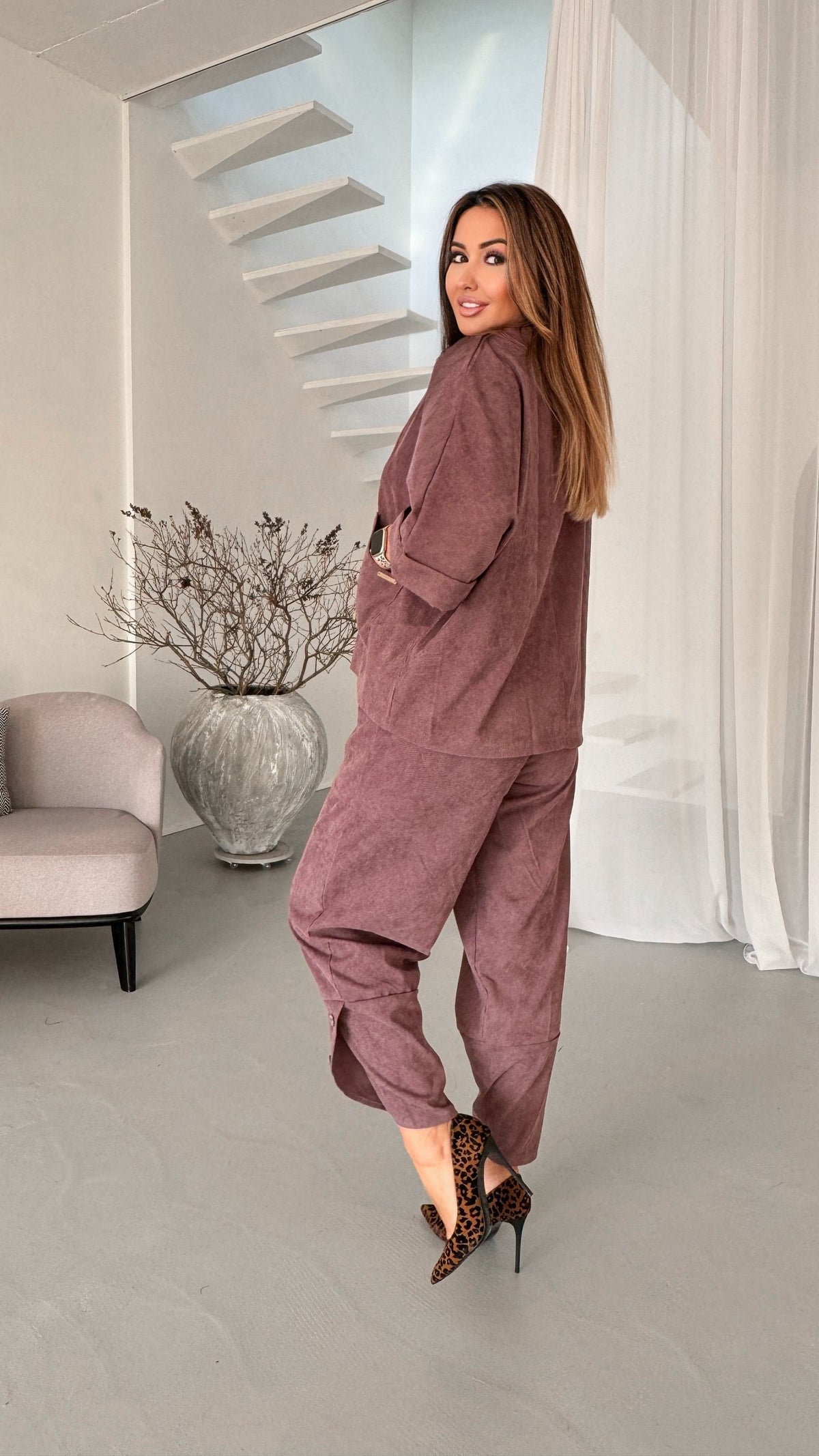 A woman modeling a matching dusty rose corduroy lounge set consisting of a long-sleeve top and wide-leg pants, styled with leopard print heels in a modern minimalist room.