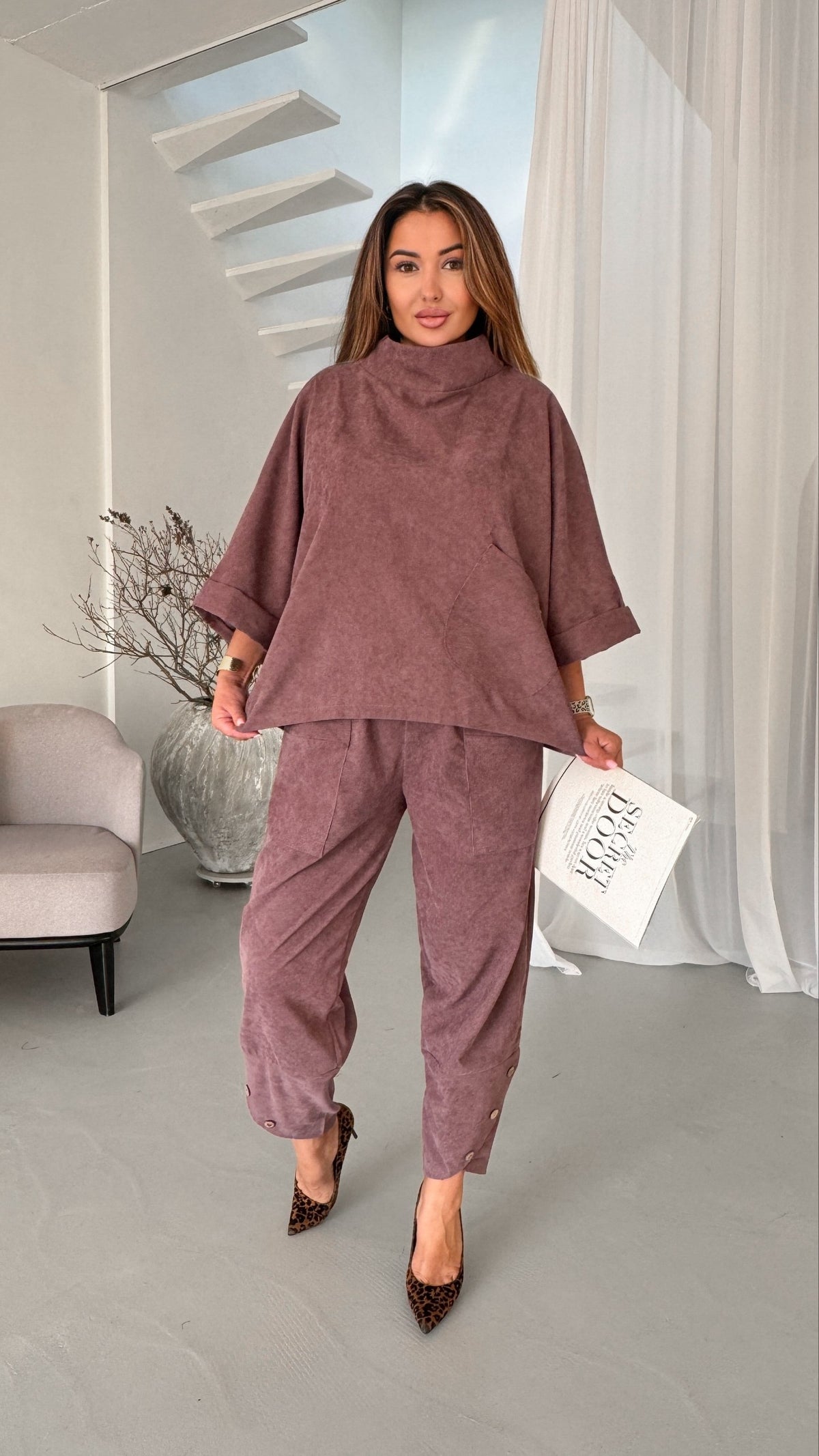 A woman models a matching mauve corduroy lounge set consisting of a high-neck, wide-sleeved tunic top and matching cropped trousers, paired with leopard print heels and holding a white magazine.