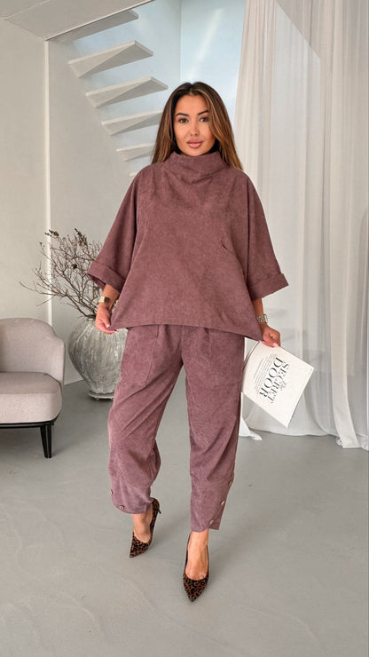 A woman models a matching mauve corduroy lounge set consisting of a high-neck, wide-sleeved tunic top and matching cropped trousers, paired with leopard print heels and holding a white magazine.