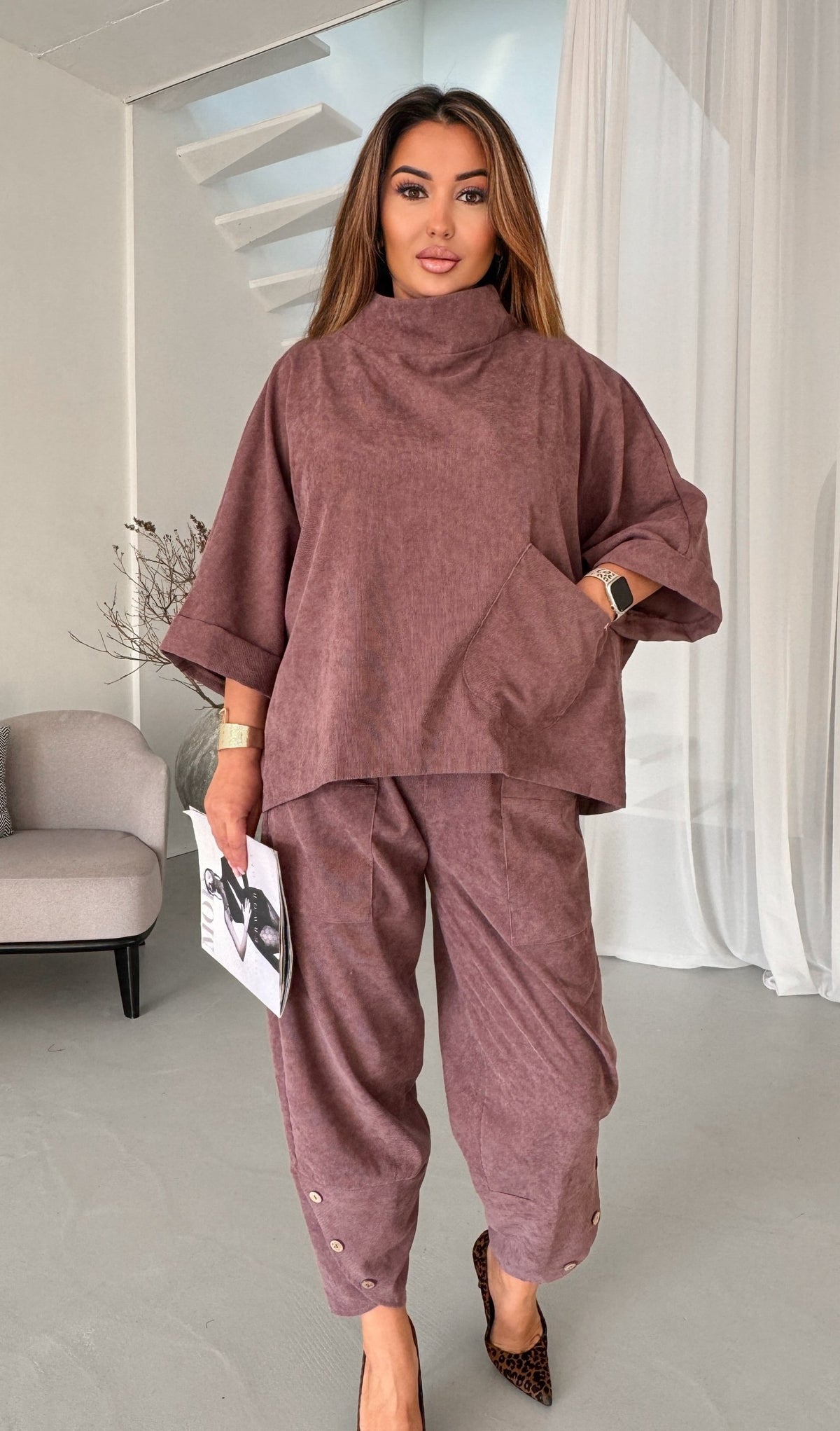 A woman modeling a matching two-piece lounge set in a dusty mauve-brown color, featuring a high-neck oversized top and matching relaxed pants, standing in a modern interior setting.