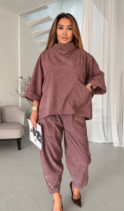A woman modeling a matching two-piece lounge set in a dusty mauve-brown color, featuring a high-neck oversized top and matching relaxed pants, standing in a modern interior setting.