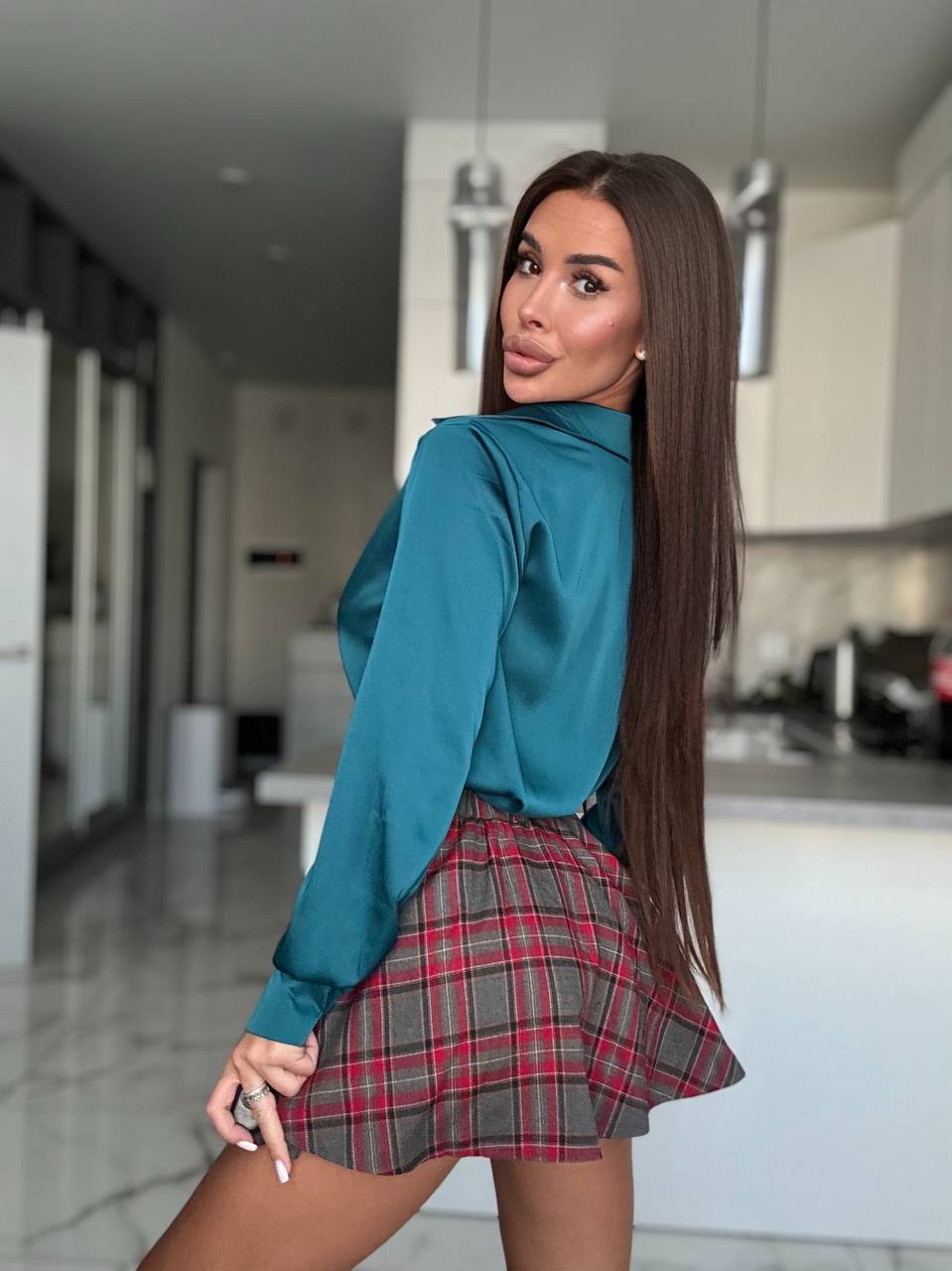 A woman posing in a modern kitchen, wearing a teal satin long-sleeve blouse and a red, gray, and black plaid pleated mini skirt.