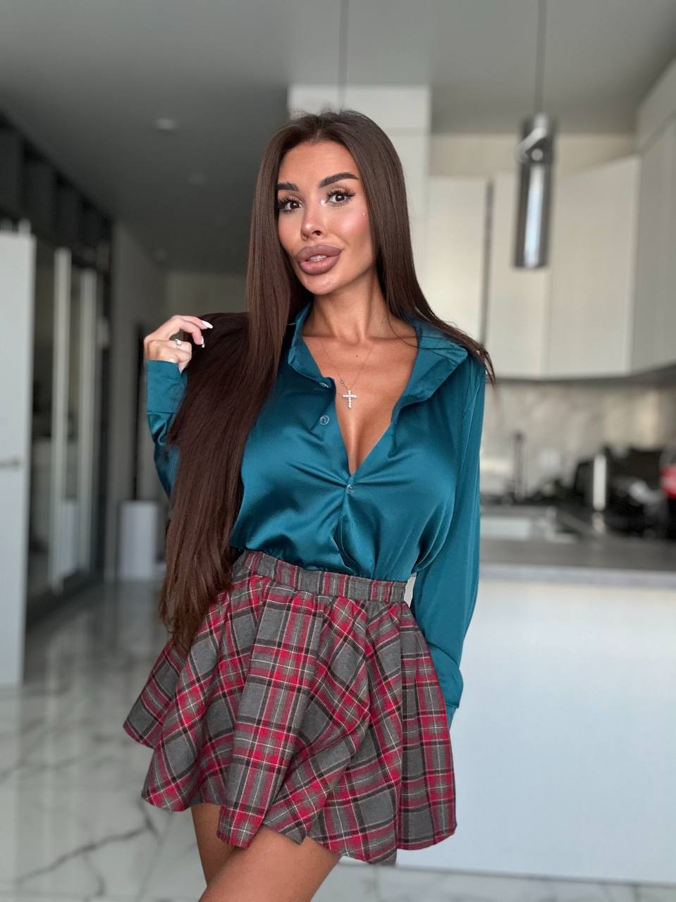 A woman posing in a modern kitchen setting, wearing a teal satin long-sleeve blouse and a red, gray, and black plaid pleated mini skirt.