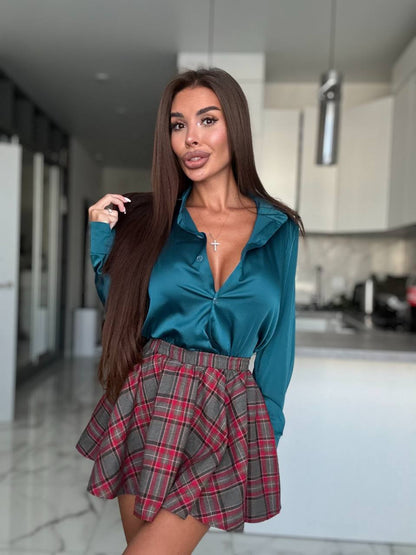A woman posing in a modern kitchen setting, wearing a teal satin long-sleeve blouse and a red, gray, and black plaid pleated mini skirt.