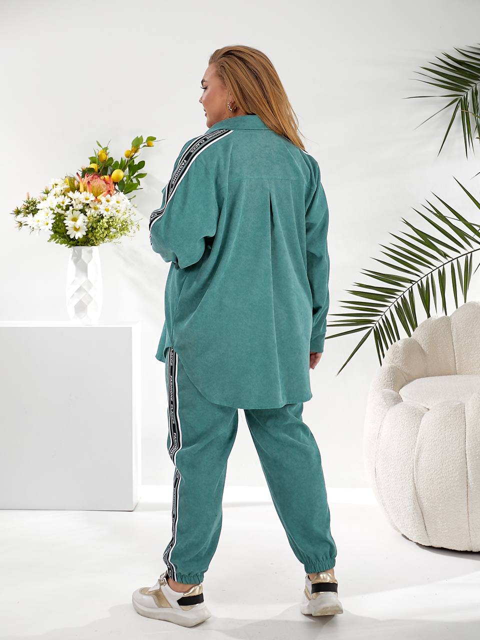 A woman wearing a matching teal green lounge set with a long-sleeve button-up shirt and jogger pants, both featuring a white stripe with black text down the side. She is shown from the back in a bright, modern room with a white pedestal, flowers, a palm leaf, and a white tufted chair.