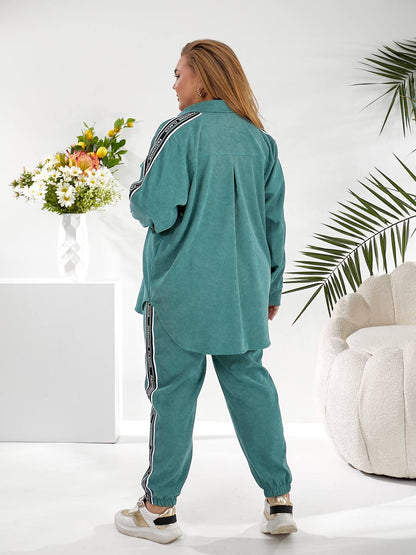 A woman wearing a matching teal green lounge set with a long-sleeve button-up shirt and jogger pants, both featuring a white stripe with black text down the side. She is shown from the back in a bright, modern room with a white pedestal, flowers, a palm leaf, and a white tufted chair.