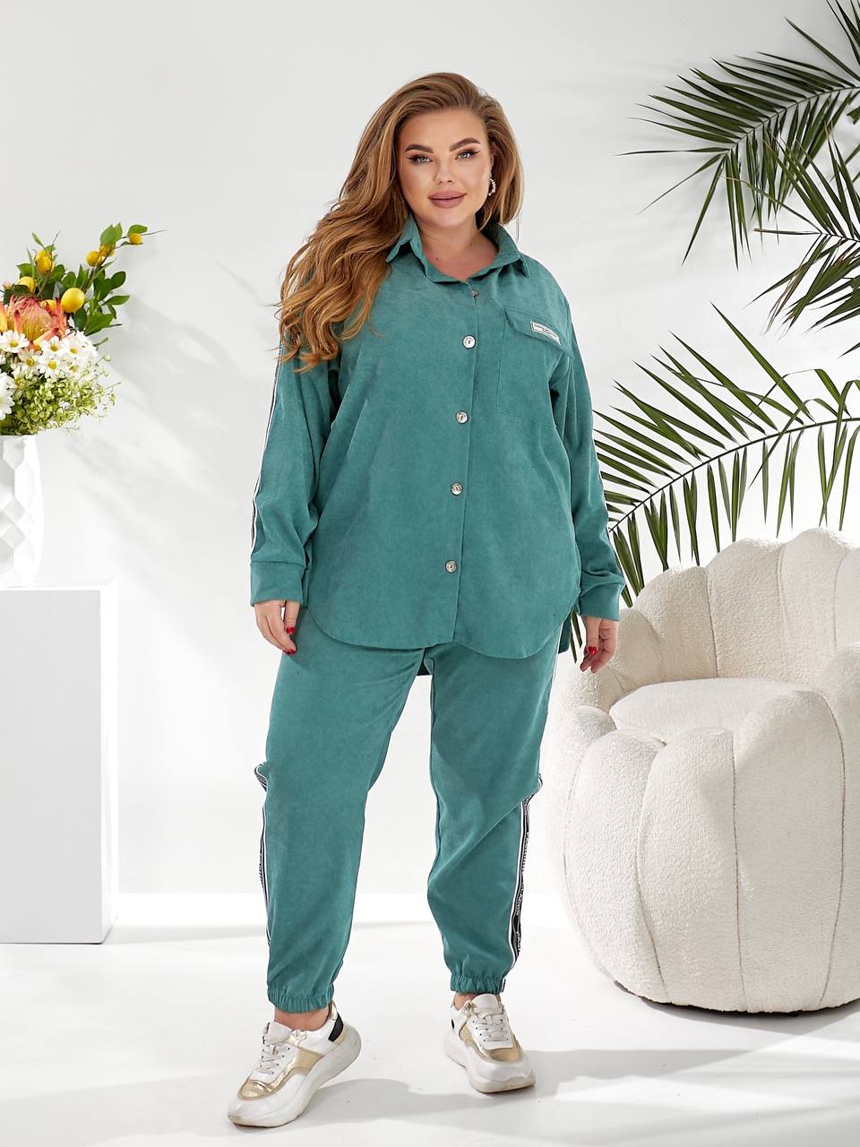 A woman modeling a matching teal lounge set consisting of a long-sleeve button-up shirt and jogger pants, styled in a bright, modern room with a white armchair and tropical plants.