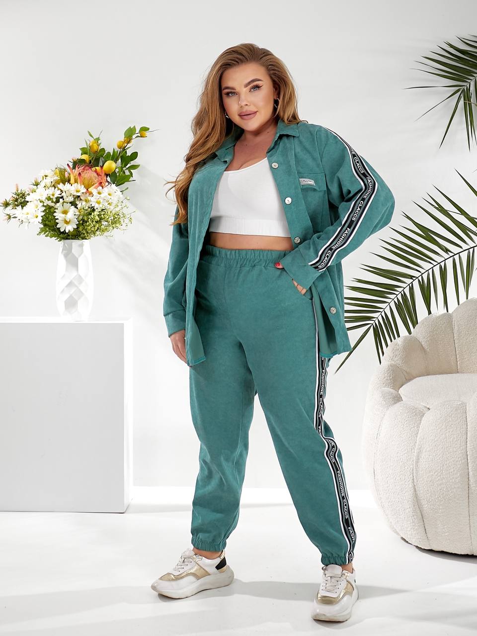 A woman modeling a matching teal green lounge set consisting of a button-up shirt and jogger pants, both featuring black and white striped side details, styled in a bright, modern room with a white armchair and a flower arrangement.