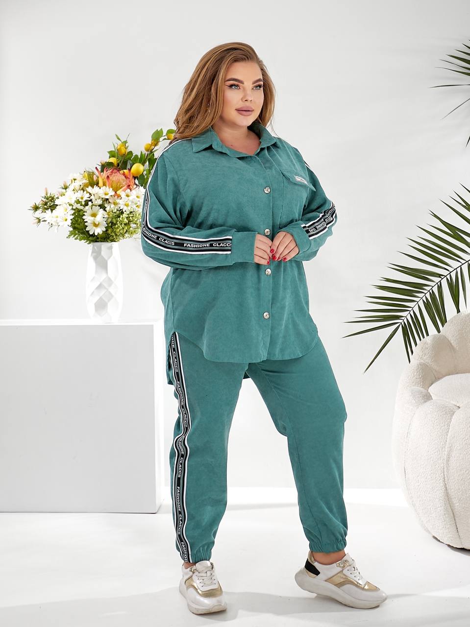 A woman modeling a matching teal green lounge set consisting of a long-sleeve button-up shirt and jogger pants, both featuring black and white striped details on the sleeves and legs, set against a bright white studio background with decorative plants and a flower arrangement.