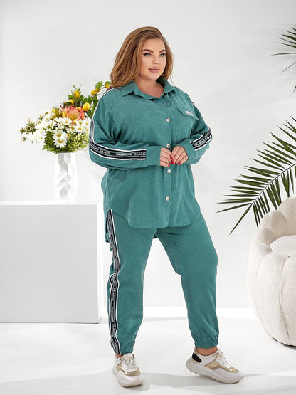 A woman modeling a matching teal green lounge set consisting of a long-sleeve button-up shirt and jogger pants, both featuring black and white striped details on the sleeves and legs, set against a bright white studio background with decorative plants and a flower arrangement.