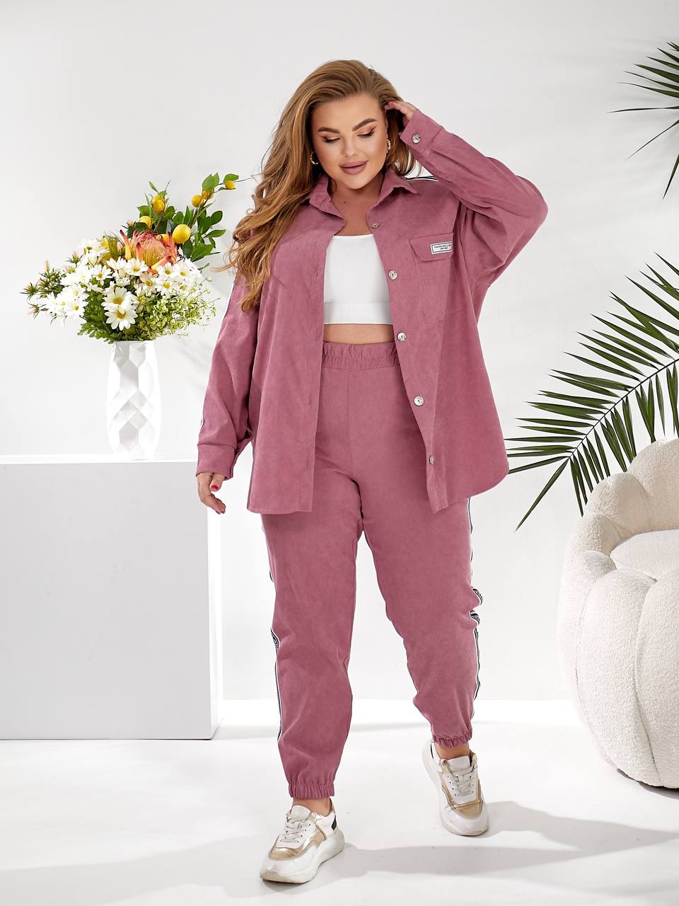 A woman modeling a dusty rose corduroy lounge set consisting of a button-up shirt and matching jogger pants, styled with a white crop top and white sneakers in a bright, modern studio setting.