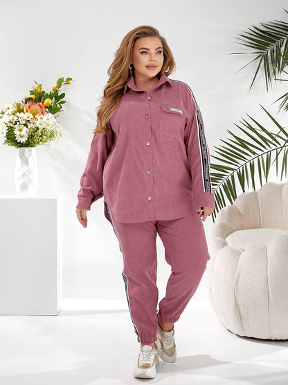 A woman modeling a matching dusty rose corduroy lounge set with black and white striped side details, standing in a bright, modern room with a white armchair, flowers, and a palm leaf.