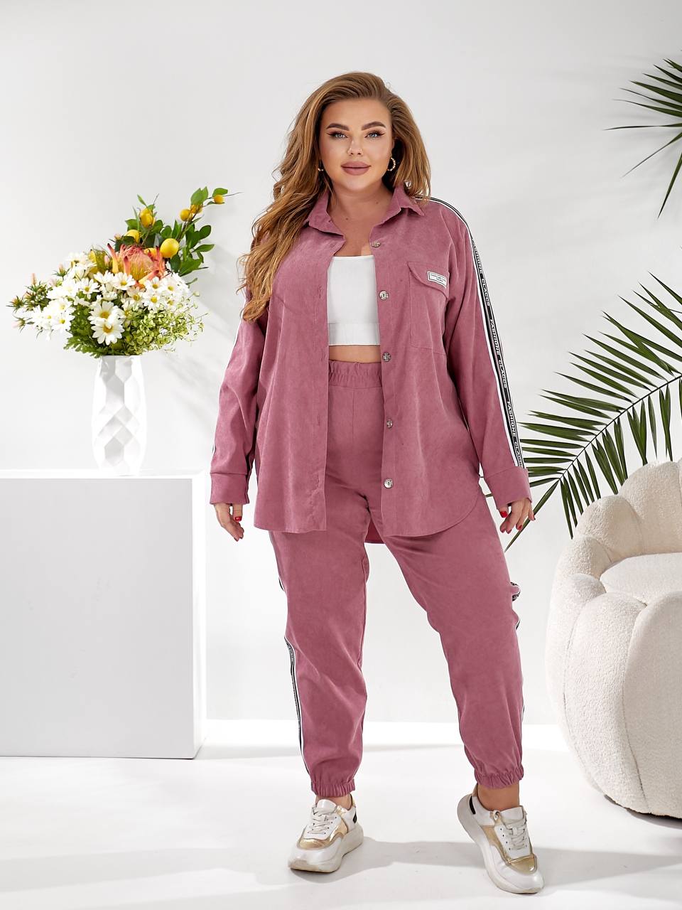A woman modeling a dusty rose two-piece lounge set consisting of a button-up shirt and matching jogger pants with white side stripes, styled with a white crop top and sneakers in a bright studio setting.