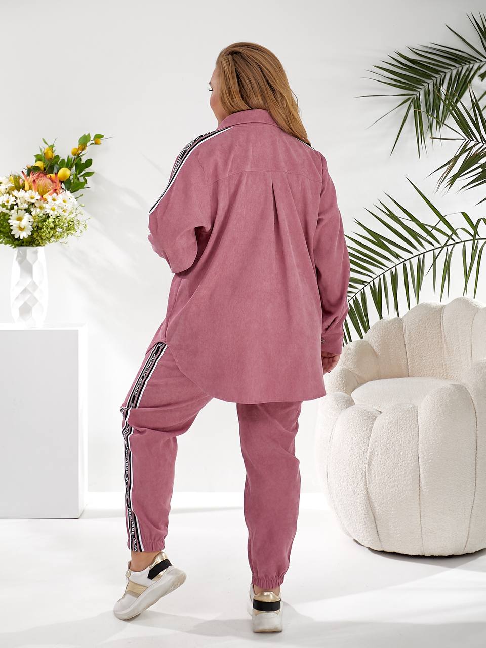 A woman wearing a dusty rose pink two-piece lounge set with black and white side stripes, shown from the back in a bright, modern room with a white armchair and a plant.