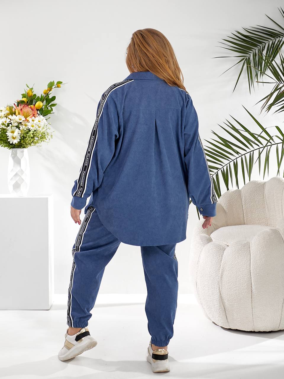 A woman shown from the back wearing a blue denim tracksuit with white stripe details on the sleeves and pants, standing in a bright, modern room with a white armchair and a flower arrangement.