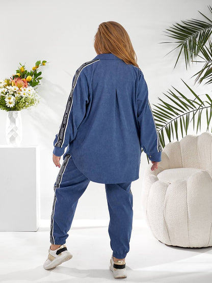 A woman shown from the back wearing a blue denim tracksuit with white stripe details on the sleeves and pants, standing in a bright, modern room with a white armchair and a flower arrangement.