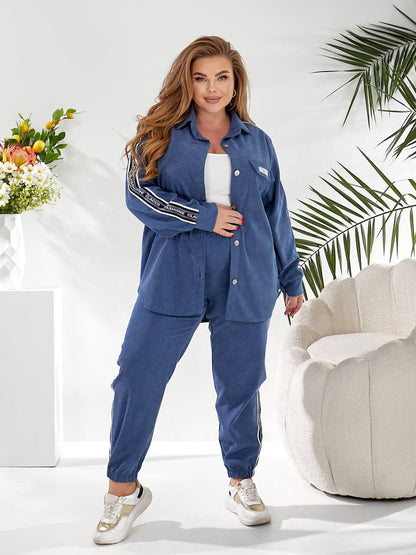 A woman modeling a matching blue lounge set consisting of a long-sleeve button-up shirt and jogger pants, styled with a white top and white sneakers in a bright, modern room with a white armchair and plants.