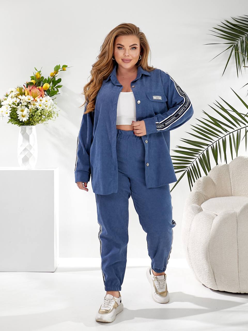 A woman modeling a matching blue lounge set consisting of a button-up shirt and jogger pants with white side stripes, styled in a bright, modern room with a white armchair and a flower arrangement.