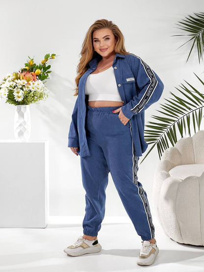 A woman modeling a matching blue lounge set consisting of a button-up shirt and jogger pants, both with white side stripes, styled in a bright, modern room with a white armchair, palm leaves, and a flower arrangement.