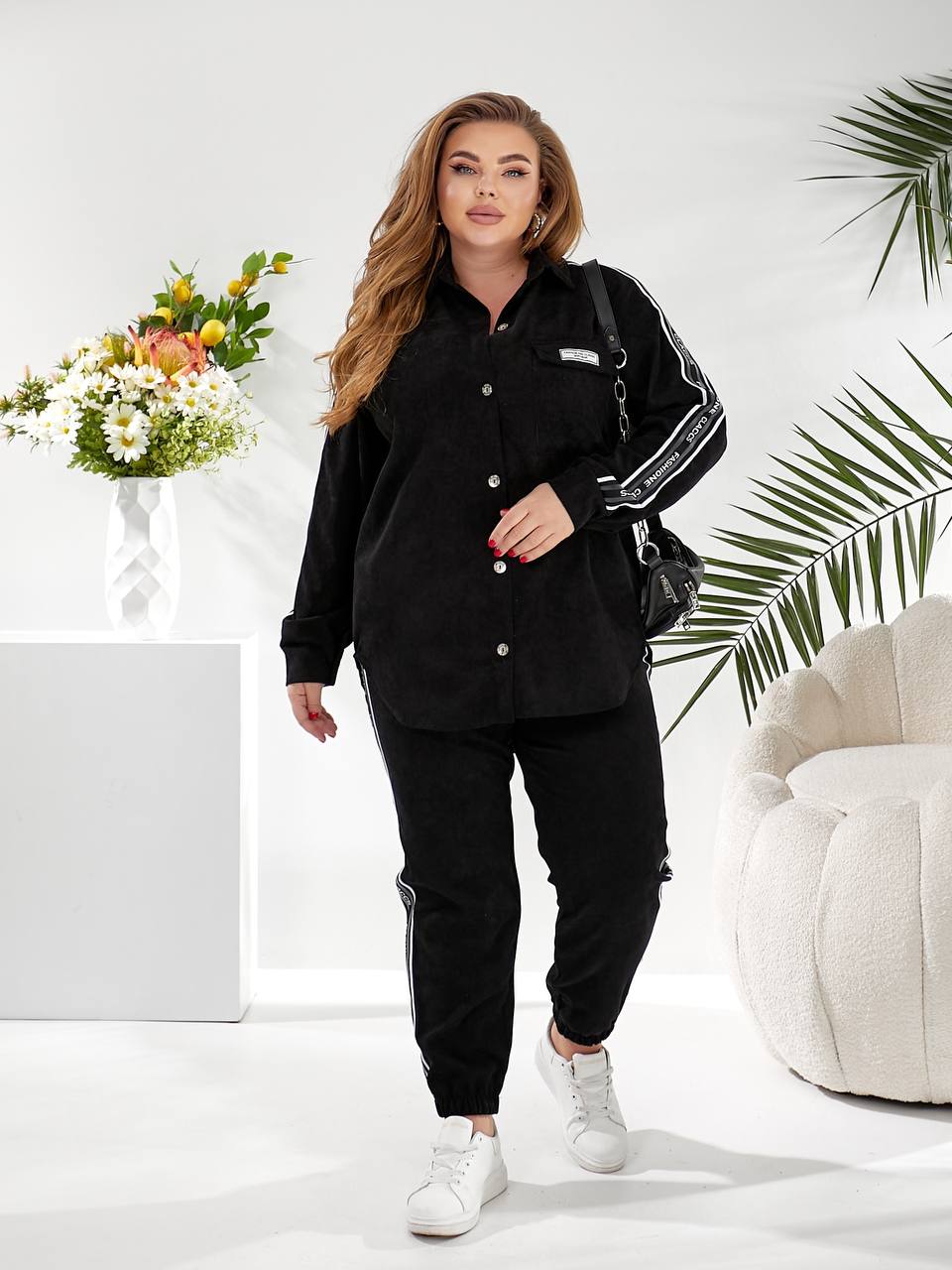 A woman modeling a black long-sleeve button-up shirt and matching black jogger pants with white side stripes, styled with white sneakers and a black shoulder bag in a bright, modern room setting.