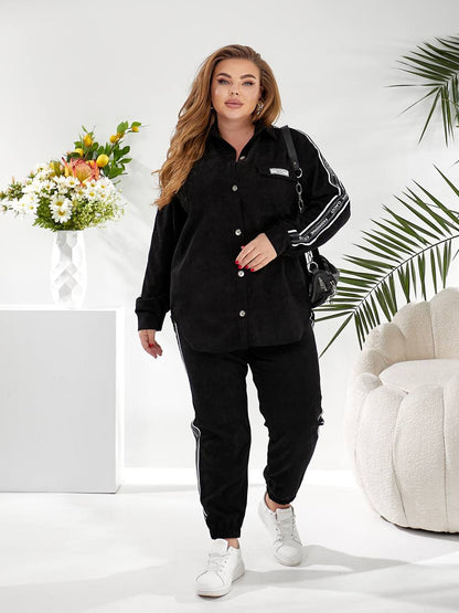 A woman modeling a black long-sleeve button-up shirt and matching black jogger pants with white side stripes, styled with white sneakers and a black shoulder bag in a bright, modern room setting.