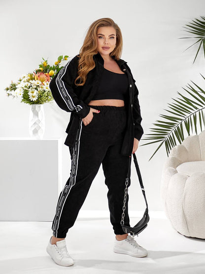 A woman modeling a black two-piece tracksuit with white side stripes, featuring a cropped tank top and matching jogger pants, styled with white sneakers and a black shoulder bag in a bright, modern room setting.