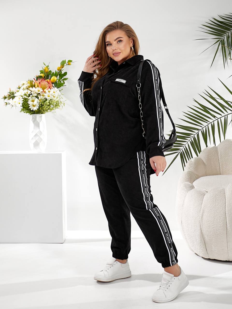 A woman modeling a black two-piece tracksuit with white side stripes, featuring a long-sleeve button-up shirt and matching jogger pants, styled with white sneakers in a bright studio setting with a floral arrangement and a white chair.
