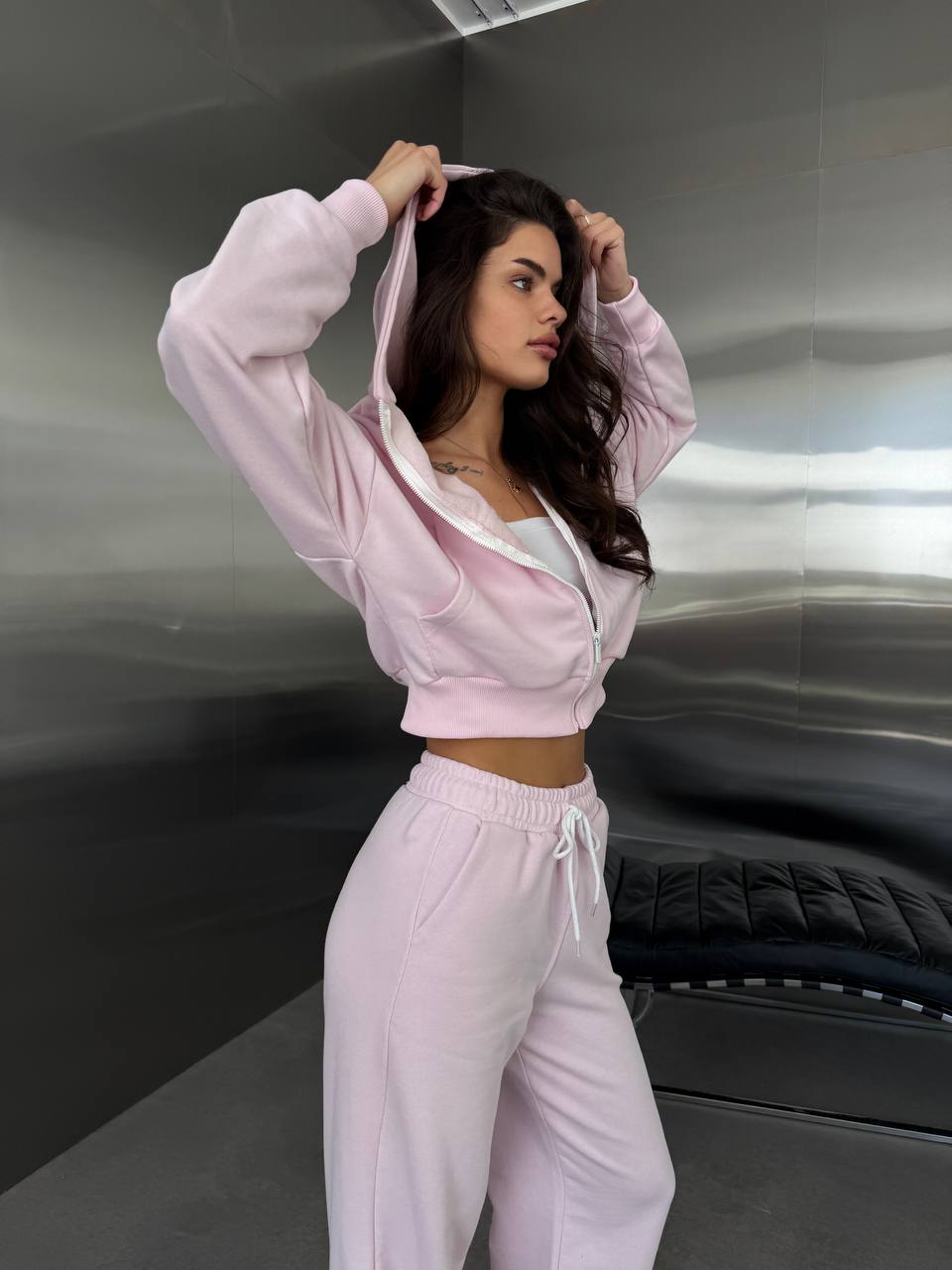 A woman modeling a light pink two-piece lounge set consisting of a cropped zip-up hoodie with white trim and matching sweatpants, standing in front of a metallic gray wall.