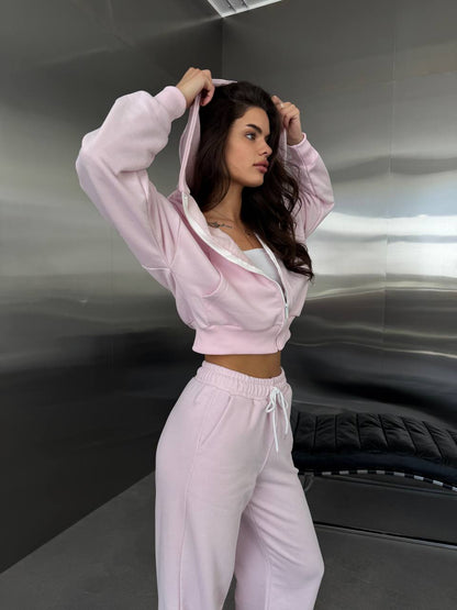 A woman modeling a light pink two-piece lounge set consisting of a cropped zip-up hoodie with white trim and matching sweatpants, standing in front of a metallic gray wall.